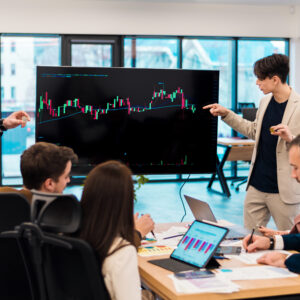 Two young workers leading business meeting in an office, discussing the topic of cryptocurrencies with other workers using a big display with currency rate and physical coins